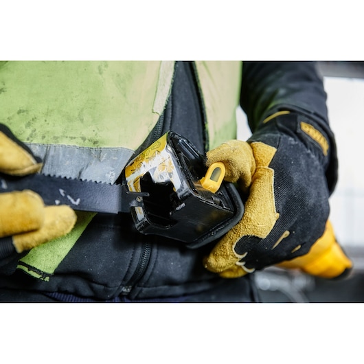 Close-up of a person wearing protective gloves and a safety vest, holding a DEWALT power tool with a blade, possibly a reciprocating saw. The tool is being operated, with focus on the hand and tool interaction.