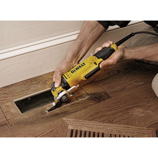 A person using a DEWALT DWE315 oscillating multi-tool to cut a section of hardwood floor near a wall.
