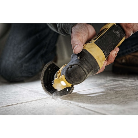 A person using a DEWALT DWE315 oscillating multi-tool to cut grout between tiles on a floor. The tool is yellow and black, and dust is visible as the cutting is performed.
