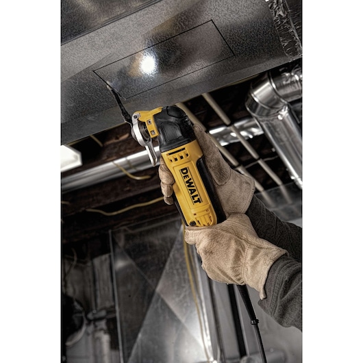 A person wearing work gloves uses a DEWALT oscillating multi-tool to cut a rectangular shape into a metal duct in an industrial setting.