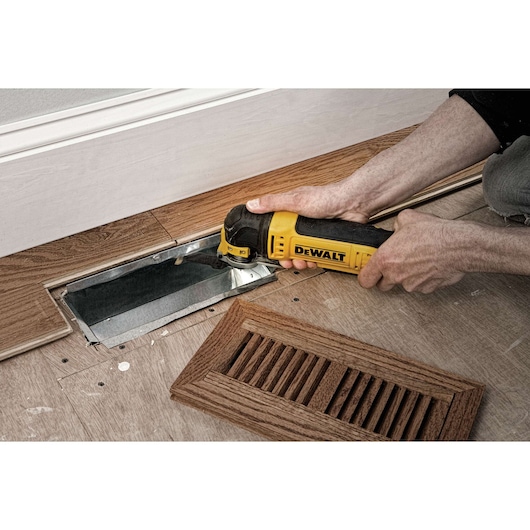 A person uses a DEWALT DWE315 oscillating multi-tool to cut wood flooring near a metal air vent opening, with a wooden vent cover placed nearby.