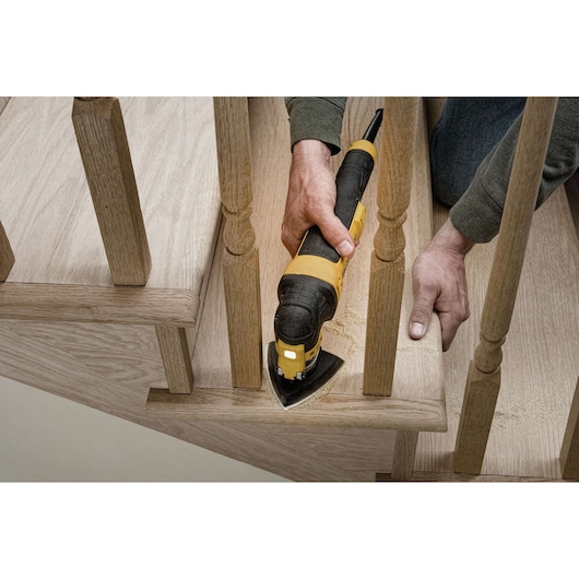 A person uses a DEWALT DWE315 oscillating multi-tool to sand or trim a wooden stair near the railing. The tool is held firmly and wood dust is visible.