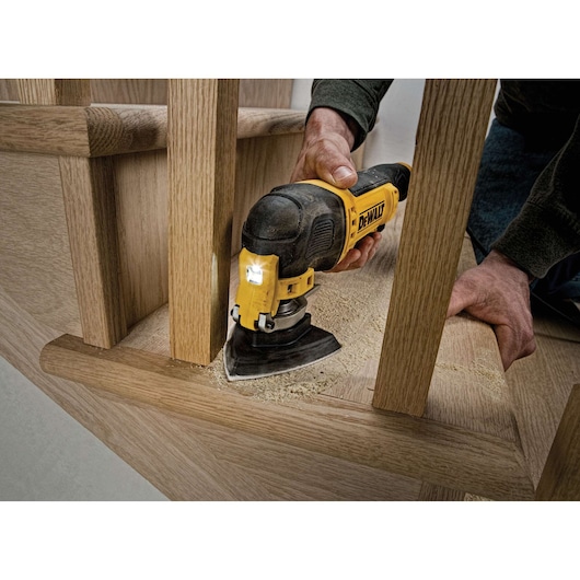 A person using a DEWALT DWE315 oscillating multi-tool to sand a wooden staircase, with sawdust visible around the tool.