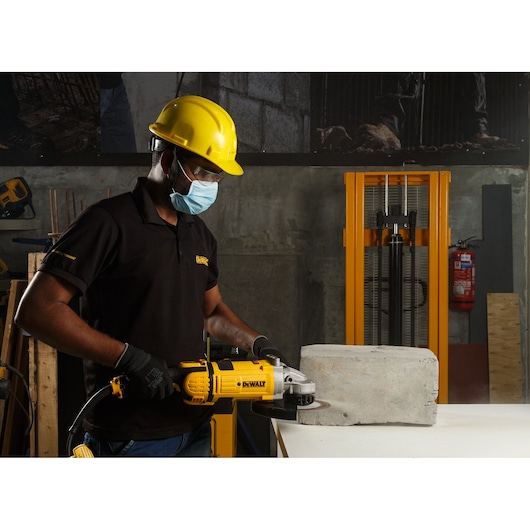 A person wearing a yellow hard hat, black gloves, and a black shirt operates a DEWALT angle grinder (model DWE4579) to cut a concrete block in an industrial workshop setting. Safety equipment and construction materials are visible in the background.