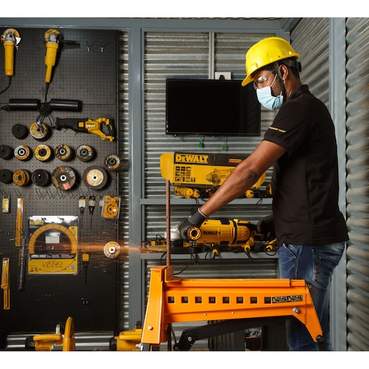 A person wearing safety gear, including a yellow hard hat, mask, gloves, and black shirt, is using a DEWALT power tool to cut a metal pipe. Sparks are flying from the cutting area. The background features a workshop with DEWALT tools and accessories organized on a pegboard and shelves.