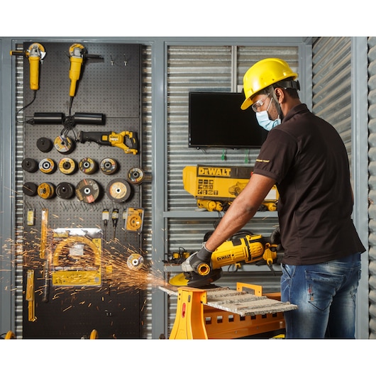 A person wearing a yellow safety helmet and black gloves is using a DEWALT angle grinder to cut metal on a workbench, with sparks flying. The background shows a wall with various DEWALT power tools and accessories organized on display.