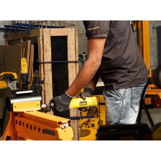 A person wearing gloves is using a DEWALT DWE4887N die grinder tool on a workpiece in a workshop. Several yellow DEWALT tools and workshop equipment are visible in the background.