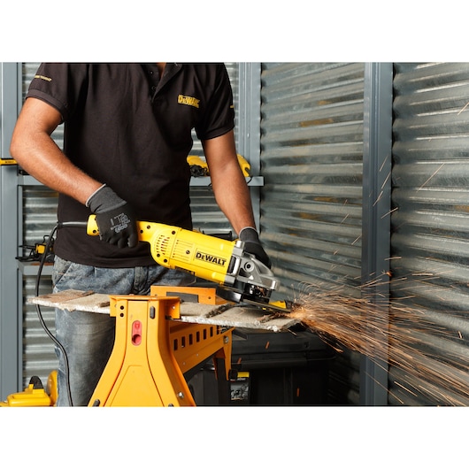 A person wearing gloves and a black shirt uses a yellow DEWALT DWE493 angle grinder to cut a metal piece, causing sparks to fly, in a workshop setting.