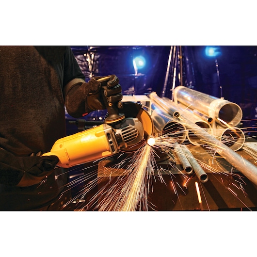 A person wearing gloves uses a yellow DEWALT angle grinder to cut metal pipes, creating bright sparks. Several metal pipes are arranged on a workbench in an industrial setting.
