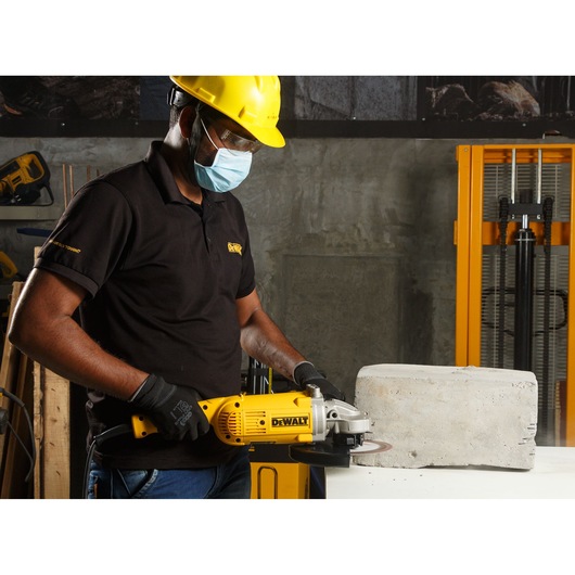A person wearing a yellow safety helmet and black gloves is using a yellow DEWALT angle grinder, model DWE496, to cut a concrete block in an indoor workshop.