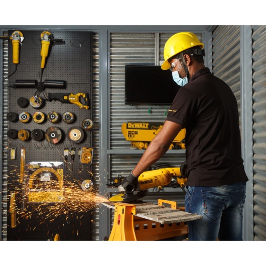 A person wearing a yellow safety helmet and black gloves is using a DEWALT DWE496 angle grinder to cut a metal piece, creating sparks. The individual is in a workshop with various DEWALT tools and accessories organized on the wall.