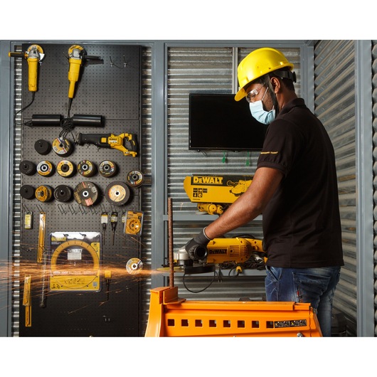 A person wearing a yellow safety helmet and black gloves operates a DEWALT angle grinder, producing sparks as they cut a metal rod in a workshop. Various DEWALT tools and accessories are organized on a pegboard wall in the background.
