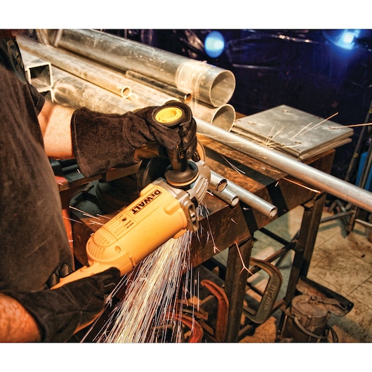 A person wearing protective gloves is using a DEWALT angle grinder to cut a metal pipe on a workbench, producing sparks. Various metal pipes and tools are visible on the workbench.