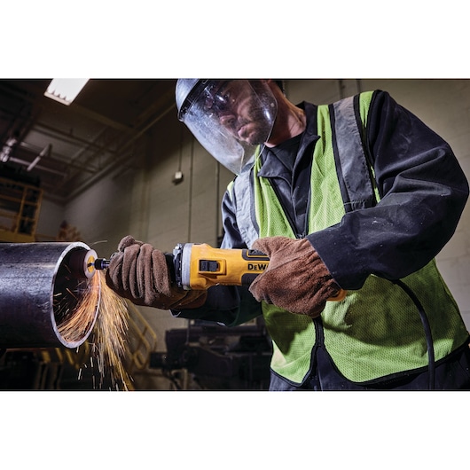 A person wearing protective gear and gloves uses a DEWALT angle grinder to cut through a large metal pipe, with sparks flying from the cutting area.