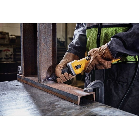 A person wearing gloves and a safety vest uses a yellow DEWALT power tool to grind or polish the edge of a metal beam in an industrial workshop setting.