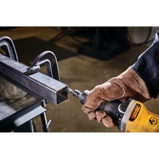 A gloved hand using a yellow DEWALT power tool to grind the edge of a metal rectangular tube, with the tube secured by clamps on a workbench.