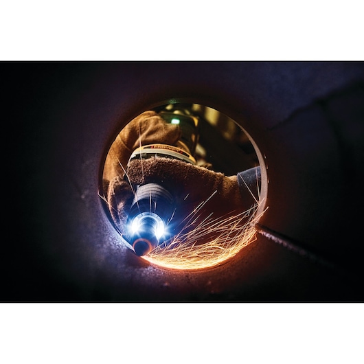 A close-up view inside a metal pipe showing a gloved hand using a DEWALT power tool, creating sparks as the tool grinds the metal.