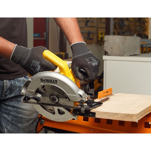A person wearing black gloves operates a DEWALT circular saw to cut a piece of wood in a workshop setting.
