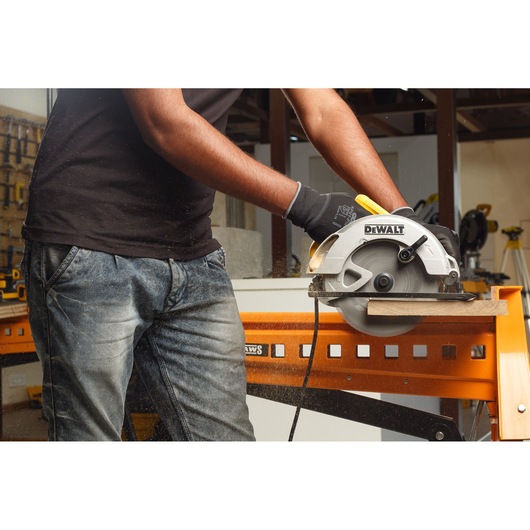 A person wearing gloves operates a DEWALT circular saw to cut a piece of wood on an orange workbench in a workshop.