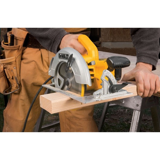 A person is using a yellow DEWALT DWE575 circular saw to cut a wooden plank. The saw is powered and positioned on a sawhorse, and the user is wearing work pants.