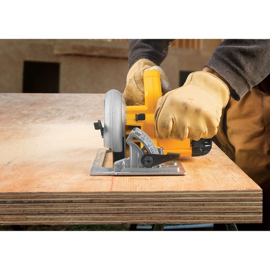 A person wearing protective gloves uses a yellow DEWALT circular saw to cut a sheet of plywood on a workbench.