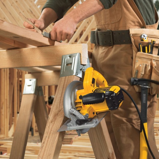 A person in work overalls is using a DEWALT DWE575 electric circular saw to cut a wooden plank on a sawhorse, with tools visible in their belt and the background showing a wood construction site.