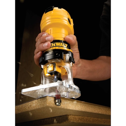 A close-up view of a person using a yellow DEWALT hand router to trim the edge of a wooden board. The router is held firmly and there are wood particles visible as it cuts.
