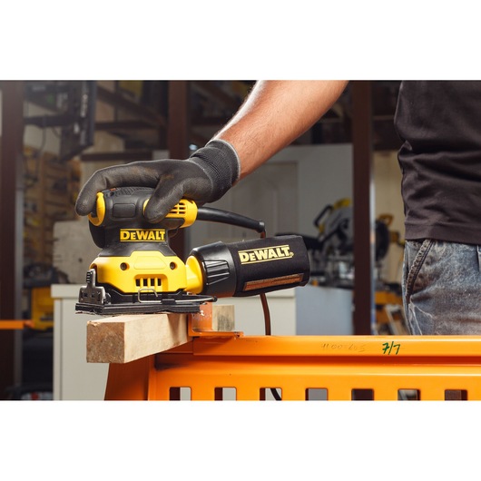 A person wearing a glove uses a DEWALT electric sander to smooth a piece of wood clamped to an orange workbench in a workshop.