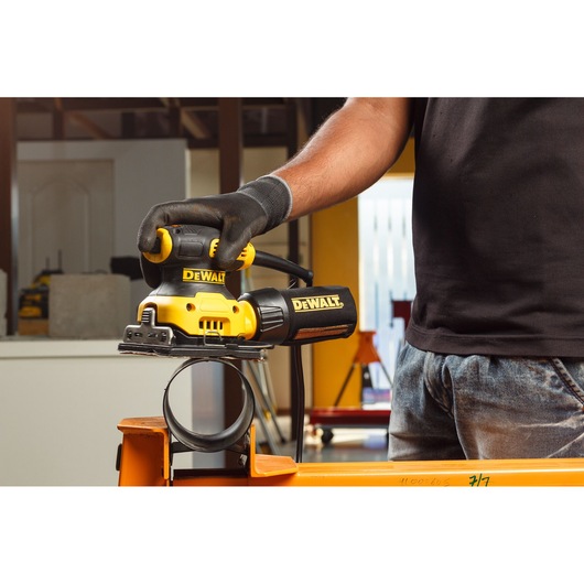 A person wearing gloves operates a DEWALT DWE6411-B5 electric sander on a metal pipe in a workshop.