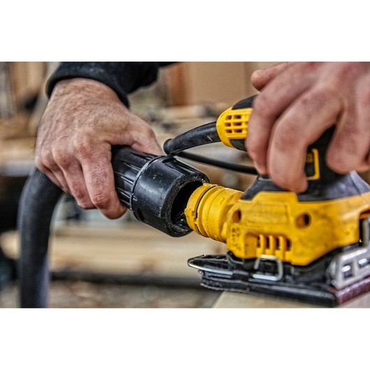 Close-up view of hands connecting a hose to a yellow DEWALT power sander, with a woodworking workspace in the background.