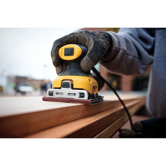 A close-up of a person using a DEWALT DWE6411 yellow and black electric sander to smooth a wooden surface outdoors. The person is wearing protective gloves.