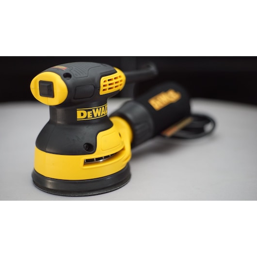 A close-up image of a DEWALT branded electric orbital sander, featuring a prominent yellow and black color scheme. The tool is positioned on a flat surface and its dust collection bag is visible in the background.