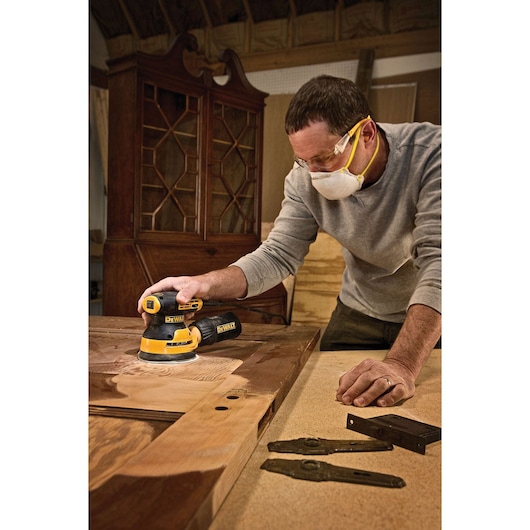 A person using a DEWALT DWE6423 random orbital sander to sand a wooden door in a woodworking shop. Some metal hardware pieces are positioned on the table nearby, and the person is wearing a protective mask.