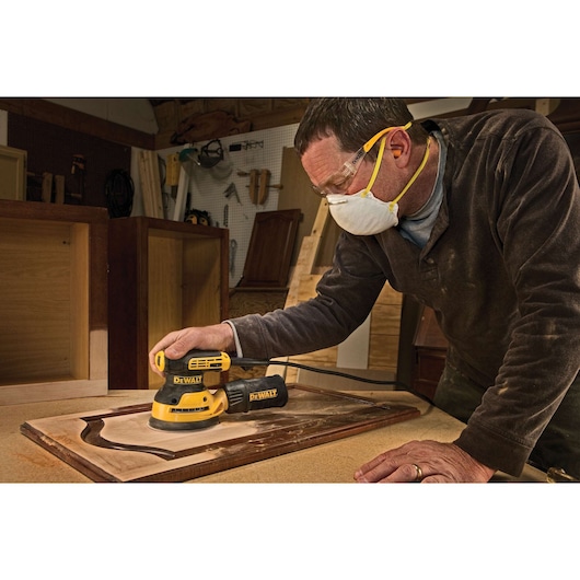 A person wearing a dust mask and safety glasses is using a DEWALT DWE6423 electric sander to smooth a wooden panel in a workshop. Various woodworking tools and cabinets are visible in the background.