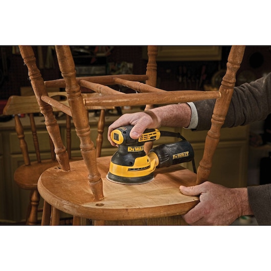 A person is using a DEWALT DWE6423 electric sander to sand a wooden chair in a workshop or garage.