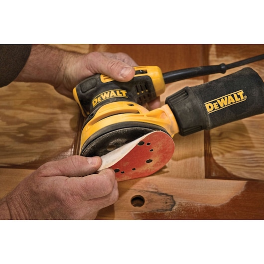 A person is changing the sandpaper pad on a DEWALT electric orbital sander, which is being used to work on a wooden surface.