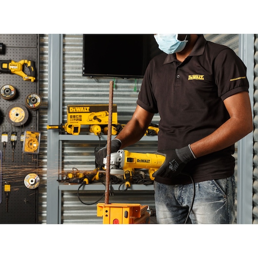 A person wearing gloves and a face mask uses a DEWALT angle grinder to cut a metal rod in a workshop. Various DEWALT tools and equipment are visible in the background.
