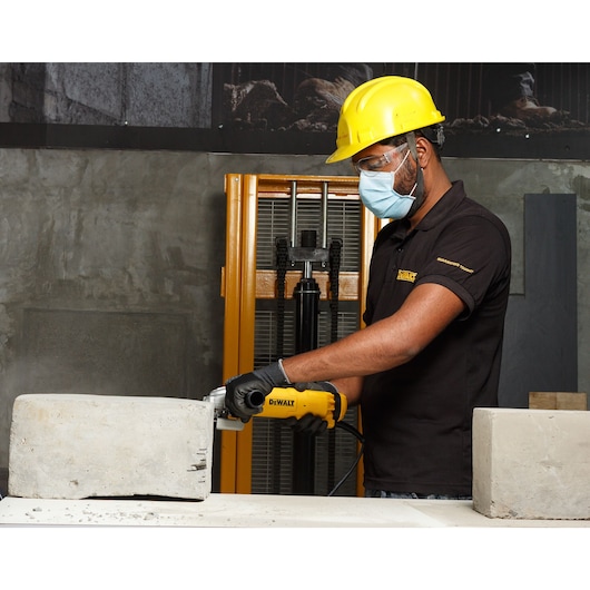 A person wearing a yellow safety helmet and gloves is using a DEWALT DWE8300S power tool to cut or grind a concrete block in an industrial environment. The person's face is blurred for privacy.