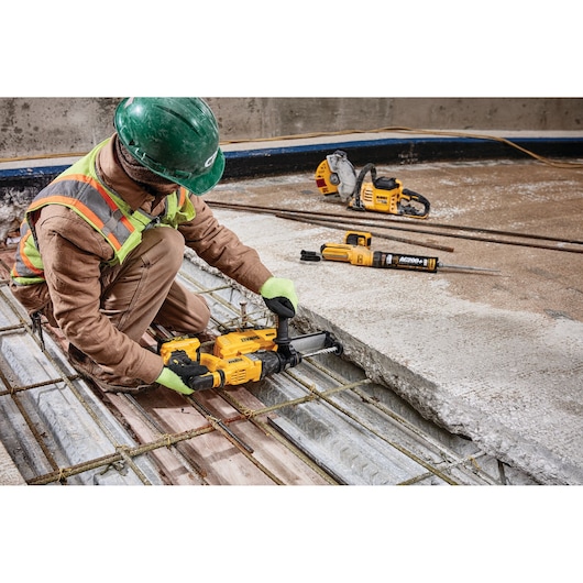 A construction worker wearing safety gear uses a yellow DEWALT power tool to drill into concrete on a worksite. Other DEWALT tools are visible on the floor nearby.