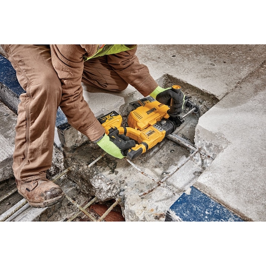 A person wearing work gloves and brown work clothes uses a yellow DEWALT cordless rotary hammer drill to break through concrete and expose metal rebar on a construction site.