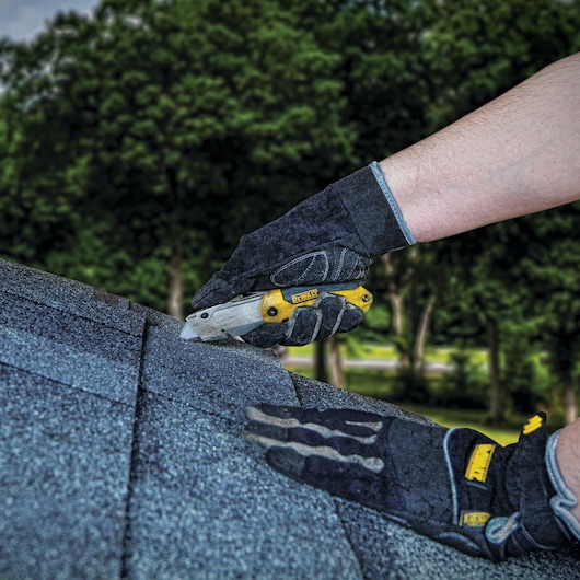 A person wearing black protective gloves is using a DEWALT utility knife to cut roofing shingles outdoors. Trees and greenery are visible in the background.