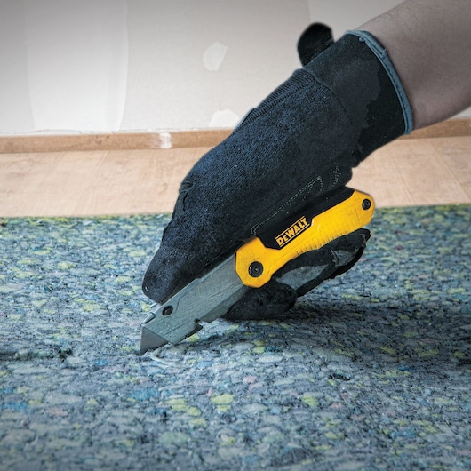 A hand wearing a black glove is using a DEWALT utility knife to cut carpet or flooring material.
