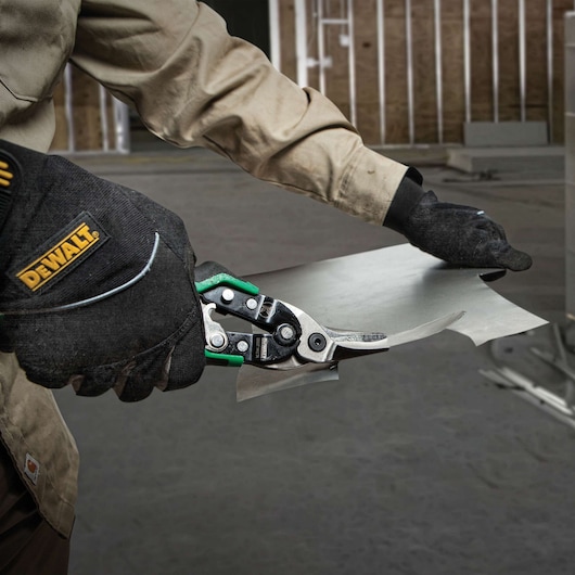 A person wearing black DEWALT gloves uses a pair of DEWALT aviation snips to cut a piece of sheet metal in an indoor workshop setting.
