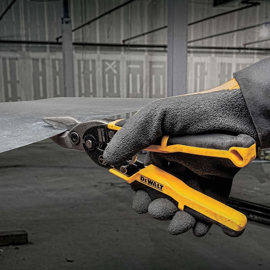A gloved hand using a DEWALT metal cutting snip to cut a sheet of metal in an industrial environment.