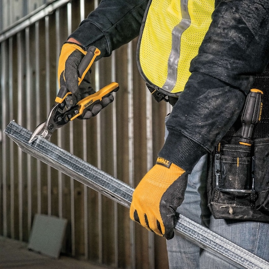 A person wearing work gloves is using a DEWALT metal snip tool to cut a metal stud in a construction setting.