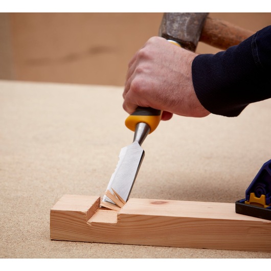 A hand uses a wood chisel with a yellow and black handle to carve a notch into a piece of light-colored wood, with a hammer striking the chisel handle.