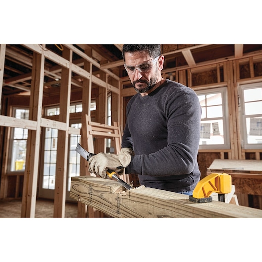 A person wearing gloves uses a DEWALT hand tool to work on a wooden beam inside a building under construction. The tool is held firmly, with wood shavings visible, and a yellow clamp secures the beam.