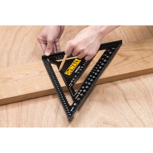 A person uses a DEWALT triangular carpenter's square (speed square) to mark a line on a wooden board. The tool has measurement markings and is placed on the board for precision.
