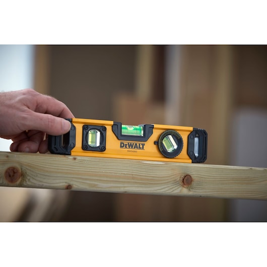 A person holding a DEWALT DWHT43003 spirit level against a wooden beam, with the tool being used to check the levelness of the surface.