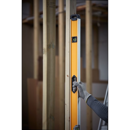 A person wearing a grey glove is holding a DEWALT spirit level against a vertical wooden beam in a construction setting.